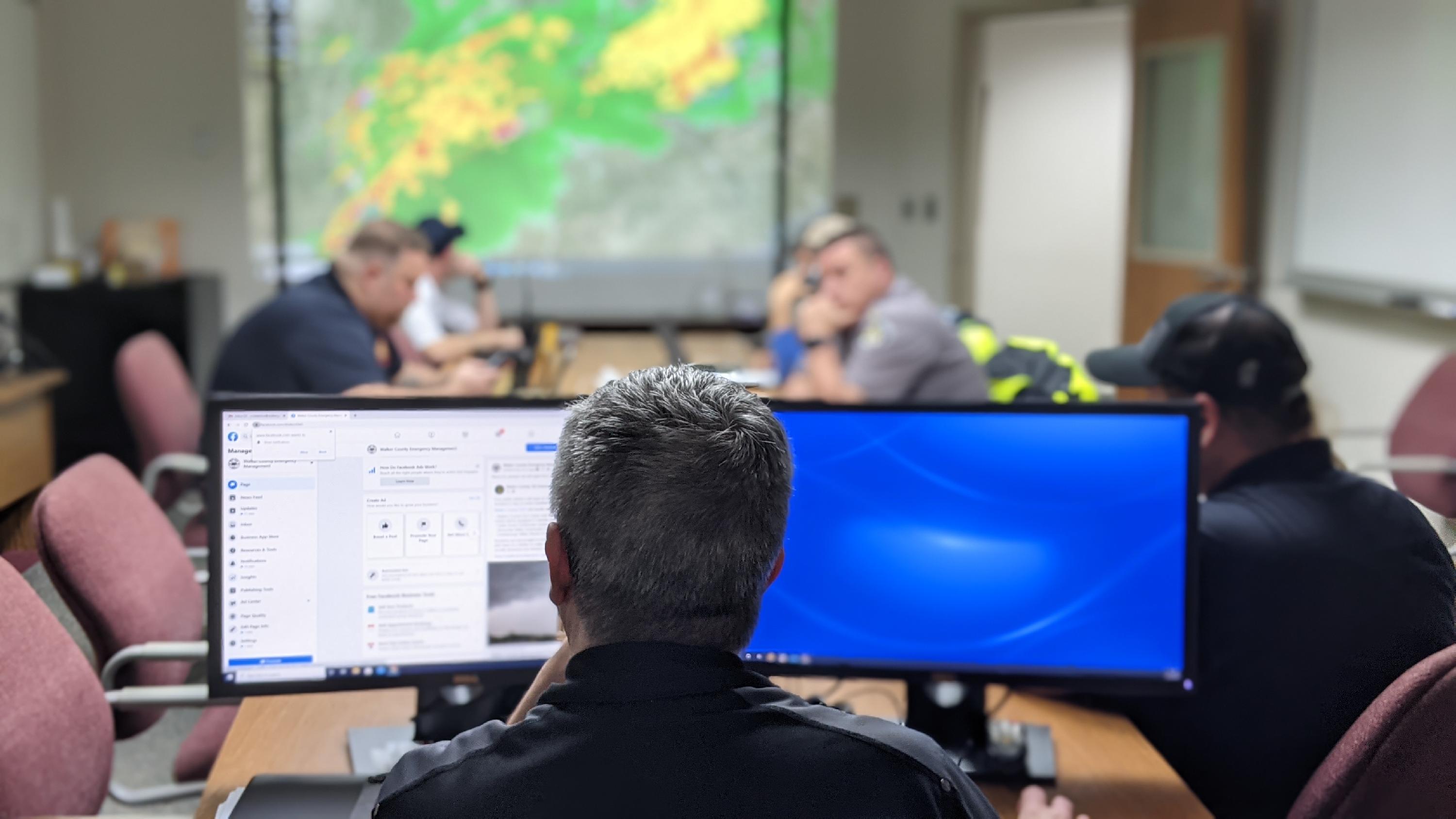 An over-the-shoulder shot from behind a man with short, graying hair as he monitors two computer screens in what appears to be an emergency operations center